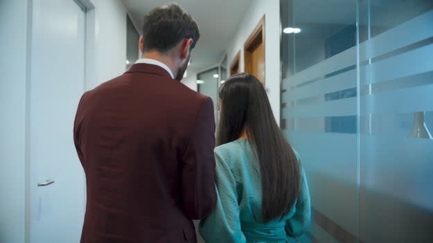 A man in a suit and a woman in a dress walk down an office hallway