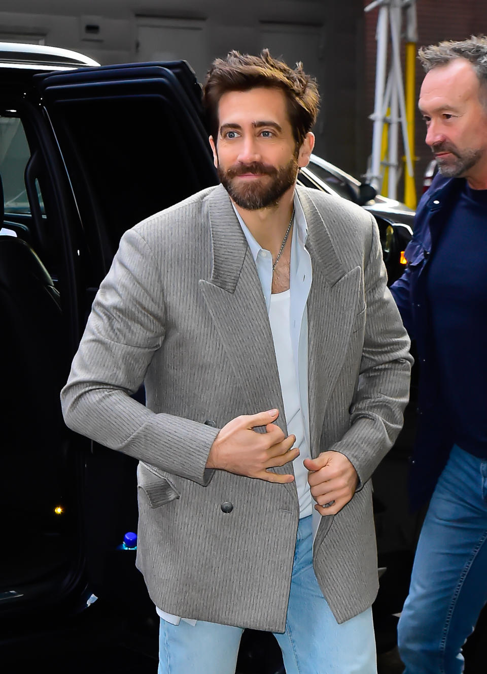 Jake Gyllenhaal smiling in a casual outfit with a blazer and jeans, exiting a car