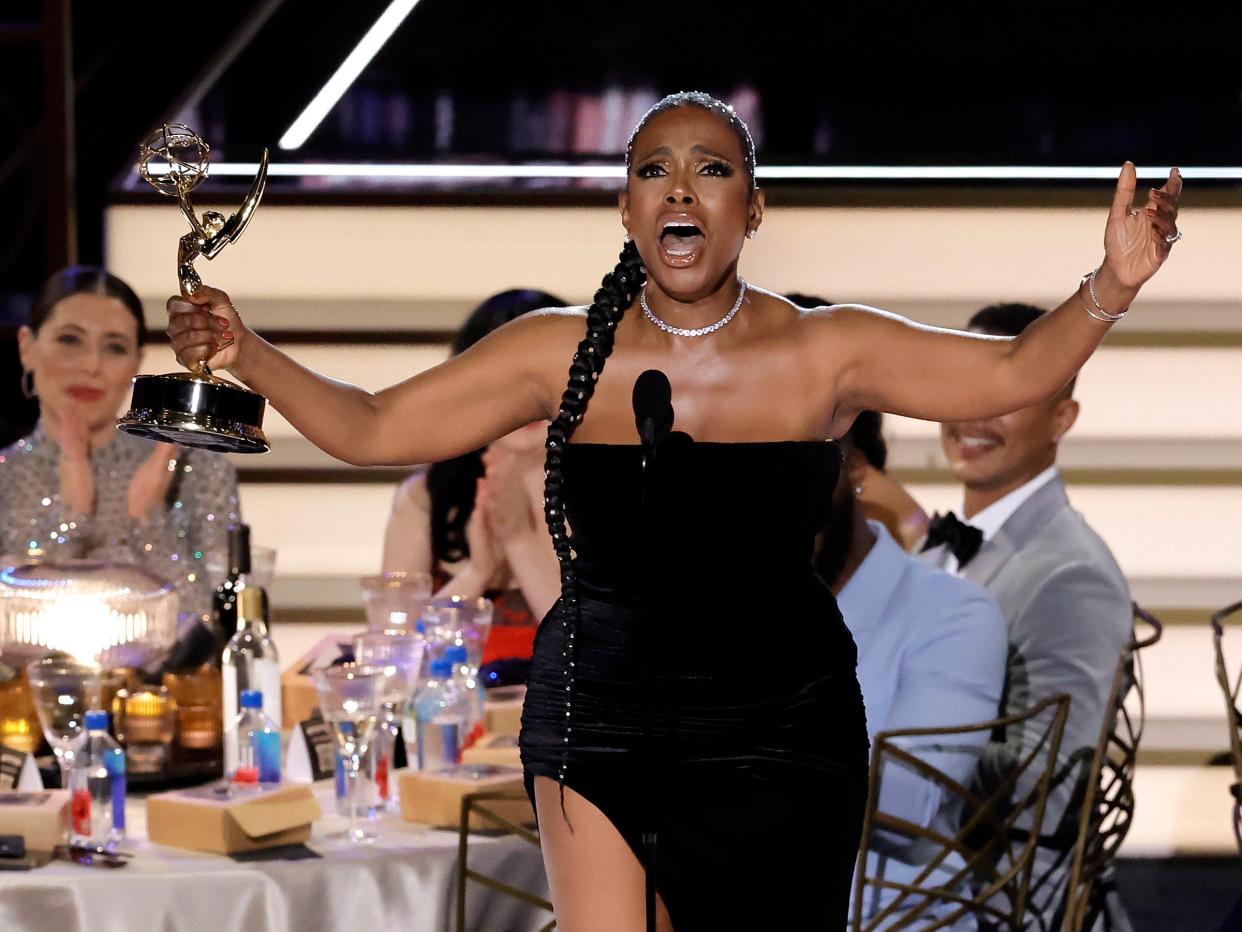 sheryl lee ralph on stage at the emmys, clad in a black dress with a slit up to her thigh and with a long brain adorned with rhinestones. she's holding an emmy in her right hand, and her arms are spread wide as she belts