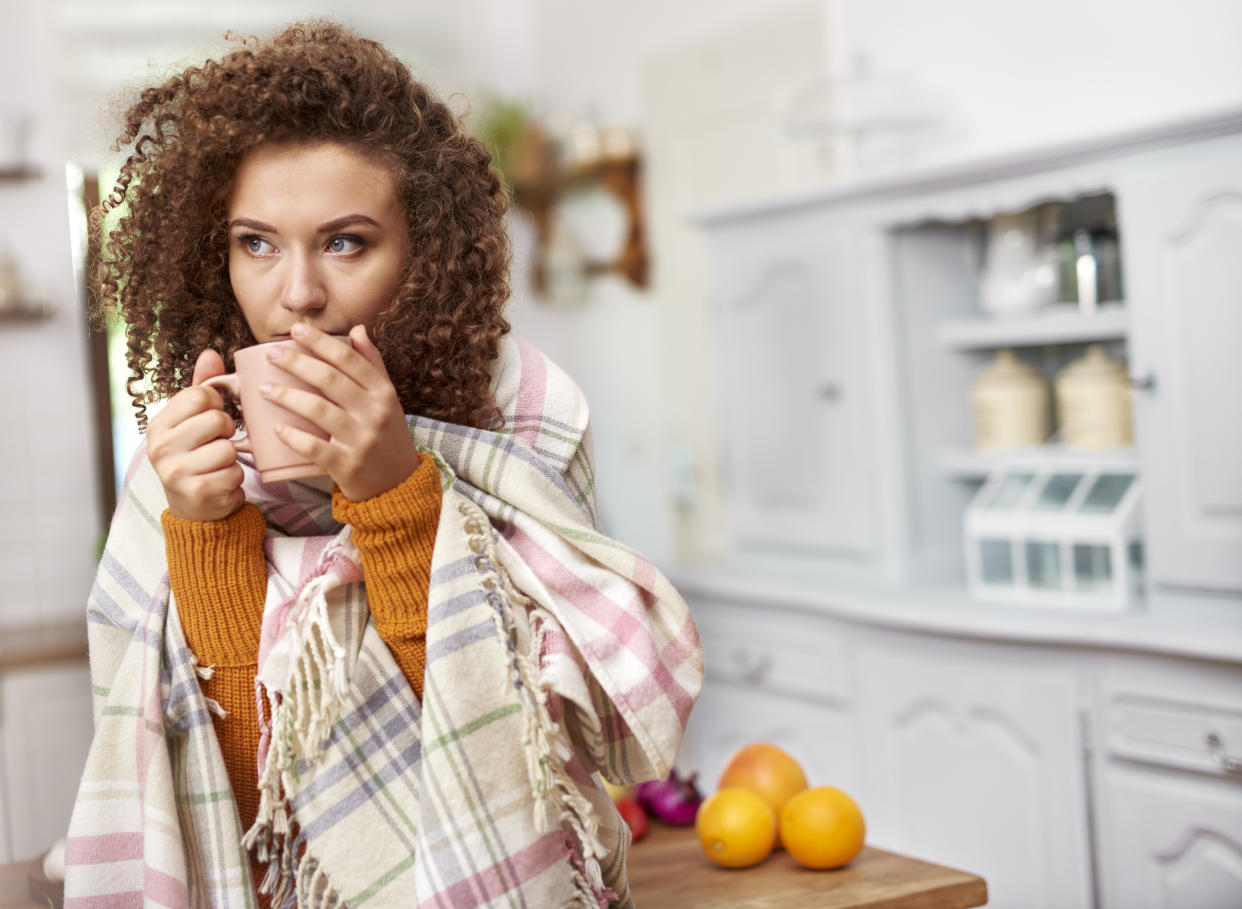 Young woman wrapped in blanket drinking hot tea to depict not putting the heating on