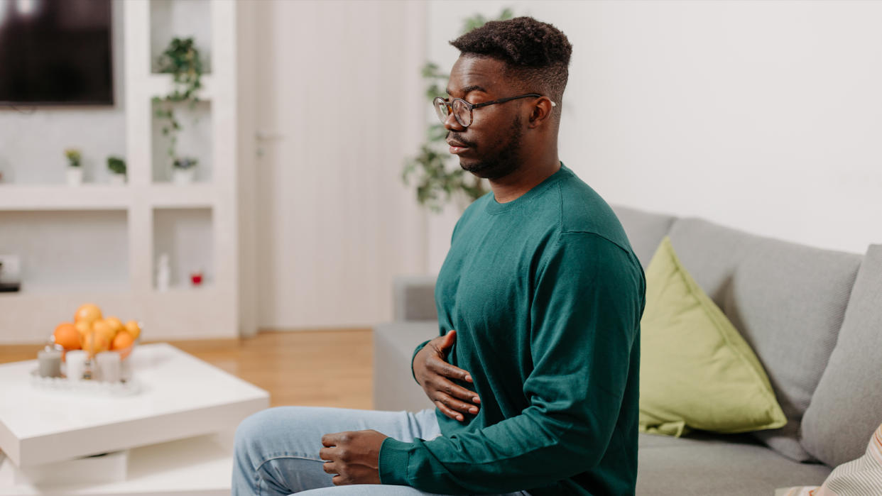  In this image, a young man is seen sitting on a sofa in his living room, looking uncomfortable and holding his stomach, presumably due to digestive problems. 