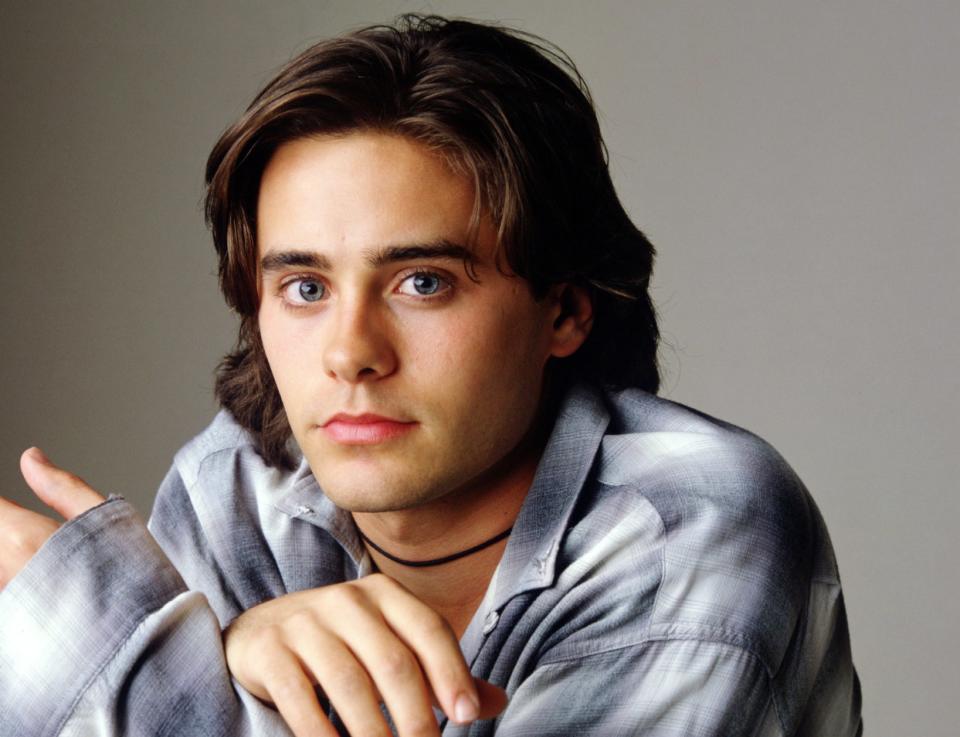 A young man rests his chin on crossed hands, wearing a casual button-up shirt, gazing forward