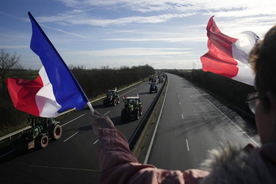 A woman cheers farmers with French flags as farmers drive their tractors on their way to a blockade on a highway, Friday, Jan. 26, 2024 near Saint-Arnoult, south of Paris. Protesting farmers shut down long stretches of some of France's major motorways on Friday, using their tractors to block and slow traffic and squeeze the government ever more tightly to cede to their demands that growing and rearing food should be made easier and more lucrative. (AP Photo/Christophe Ena)