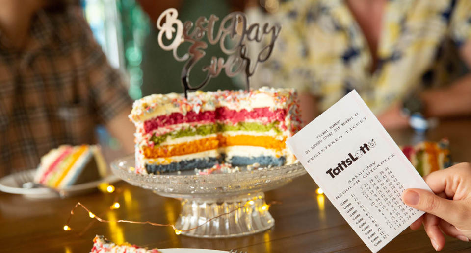 A picture of a rainbow cake with a TattsLotto ticket held in front of it.
