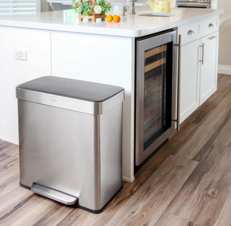 Stainless steel trash can with foot pedal next to a kitchen cabinet and under-counter oven