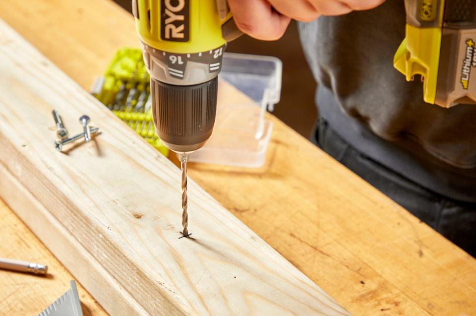 Man points drill bit into an X on a wood plank.