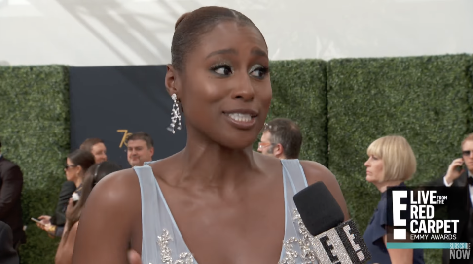Issa Rae stands on a red carpet, wearing a blue sleeveless gown and long earrings, there's a microphone in front of her