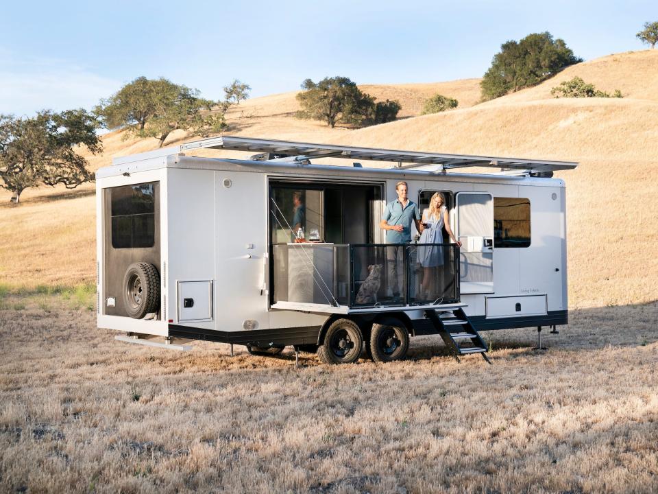 The exterior of the travel trailer as it sits on a brown field. The patio is extended and people are standing on it.