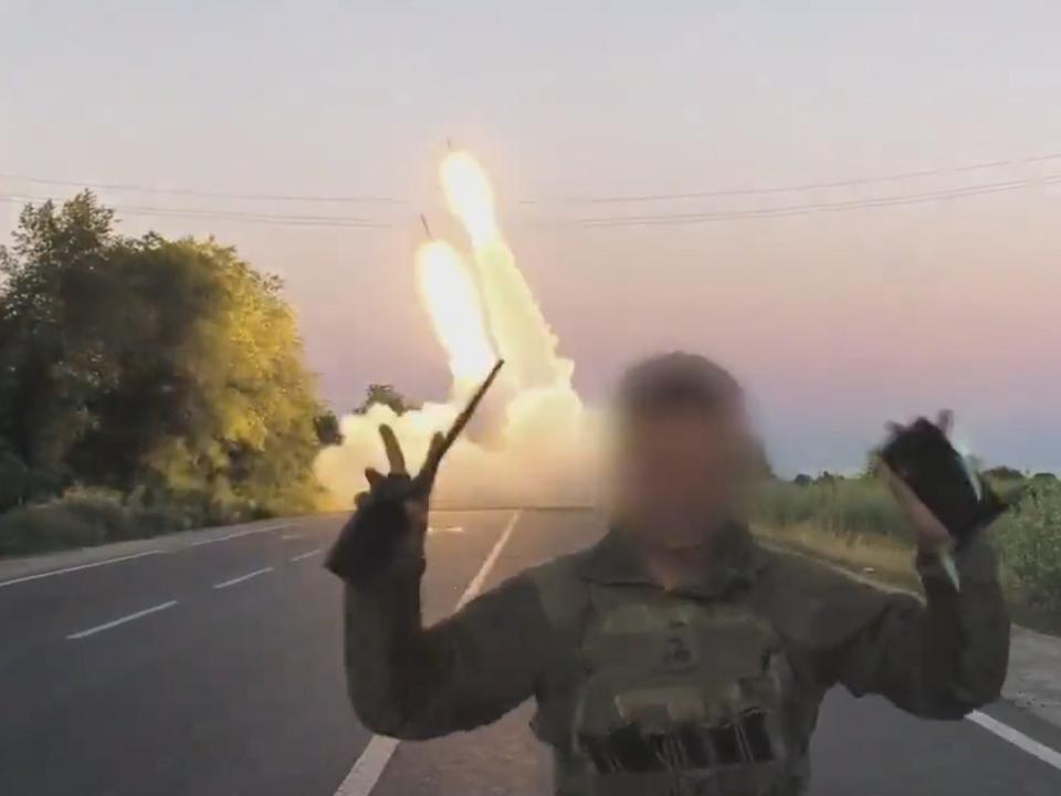A still from footage shared by the Ukrainian Defense Ministry shows a soldier, whose face is obscured, raise his hands in a "v for victory" as a US-donated HIMARS system launches rockets in the background.