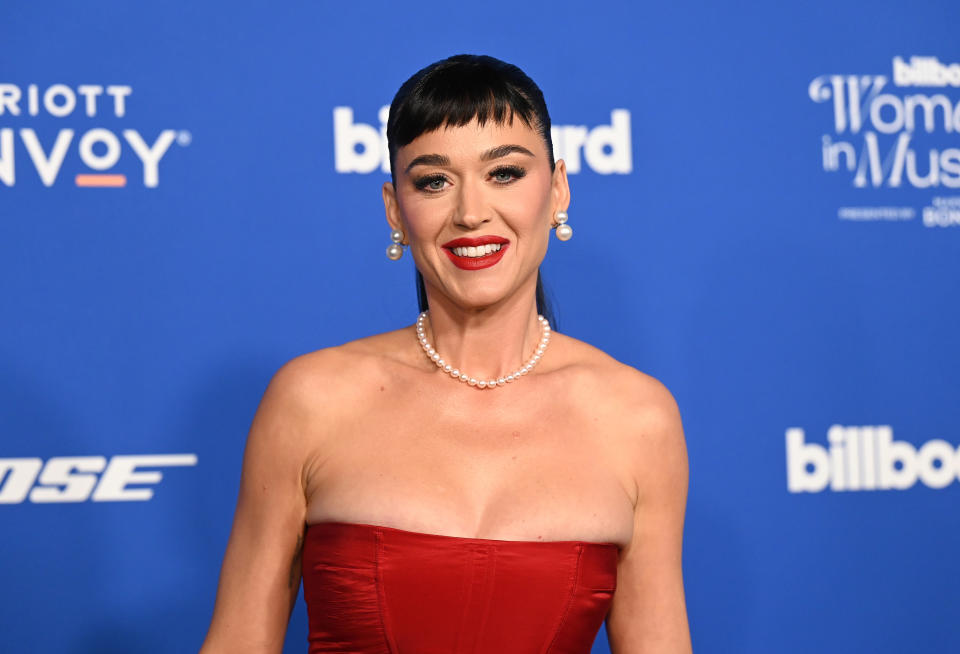 Katy smiling in a red strapless gown with a pearl necklace at a media event
