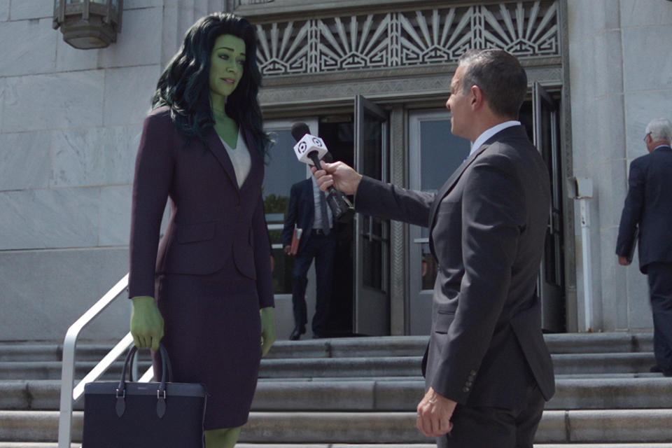 A tall female lawyer with green skin wearin g a purple suit and speaking to a reporter with a microphone on the steps outside a courthouse; still from "She-Hulk: Attorney At Law."