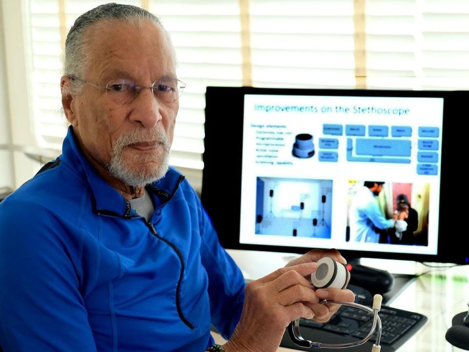 james west sits at a desk and looks at the camera, he holds a circular device in his hands, he wears a bright blue quarter zip top and glasses, behind him is a computer and window blinds