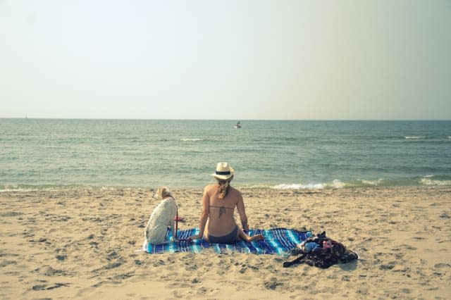 Young woman sitting on the beach with her dog, she is dressed in bikini and straw hat and watching relaxed the boat in the sea,