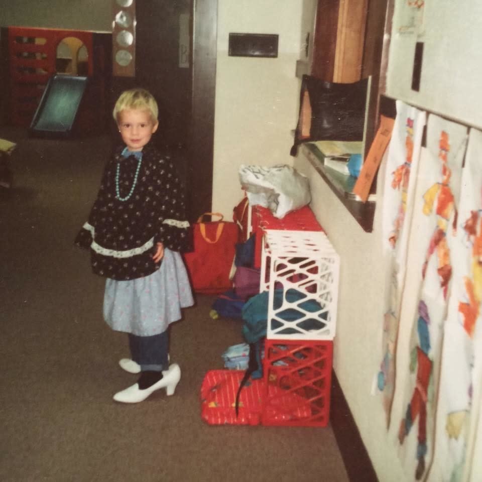 A young child with short hair wearing women's high-heel shoes, a skirt, and a necklace