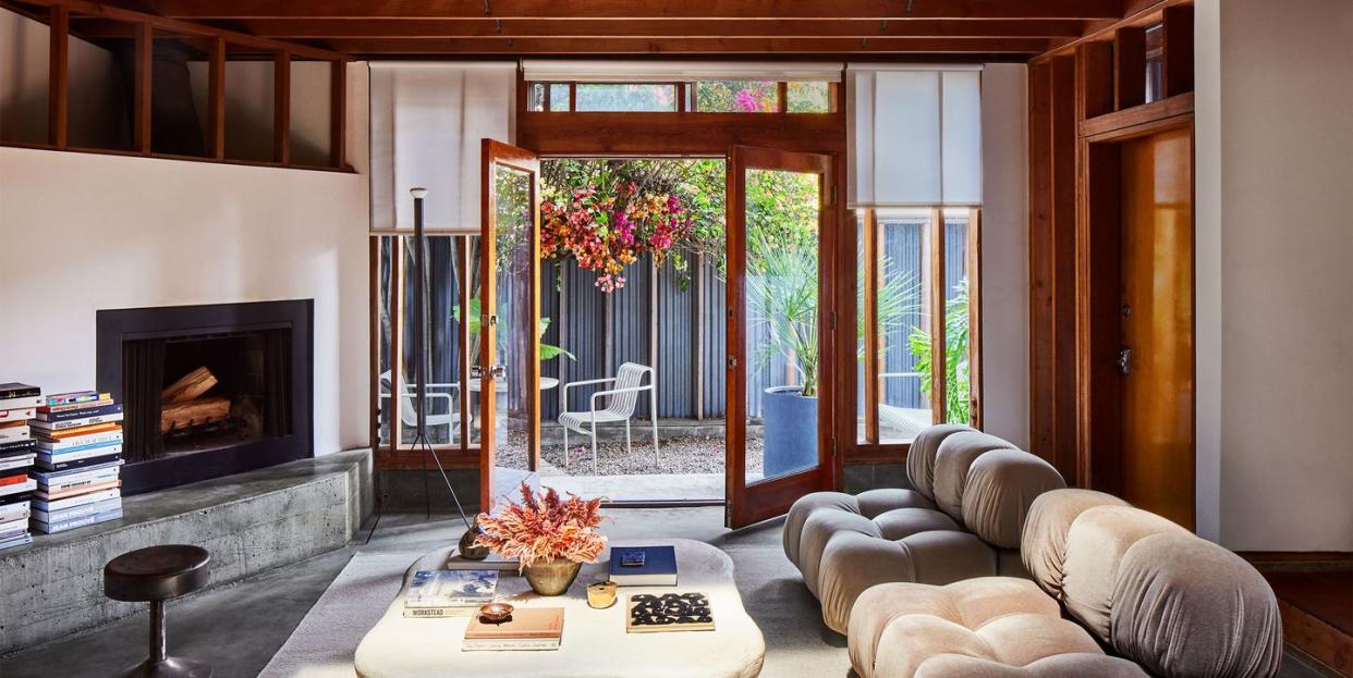 living room with small black fireplace and marble bench in front, twin greige fabric sofas and a low white plaster cocktail table, gray rug, stained woodwork and ceiling beams, doors opening to patio