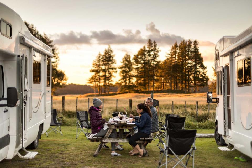Man and two women eating dinner outdoors around a wooden picnic table as the sun sets