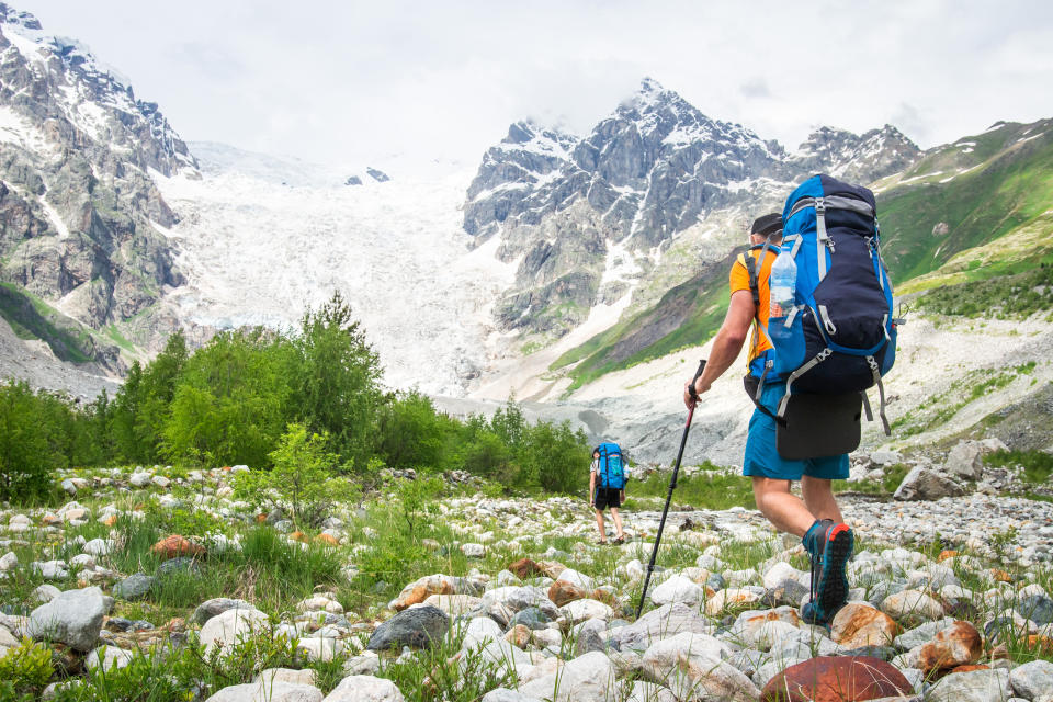 Two hikers with large backpacks and trekking poles walk across a rocky terrain towards snow-covered mountains