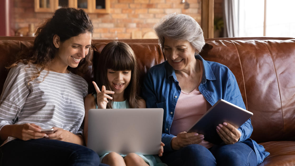 Happy kid girl, young mother, older grandmother using digital devices, laptop, tablet, making video call, watching movie online, reading book. Three family female generations using electronic gadgets
