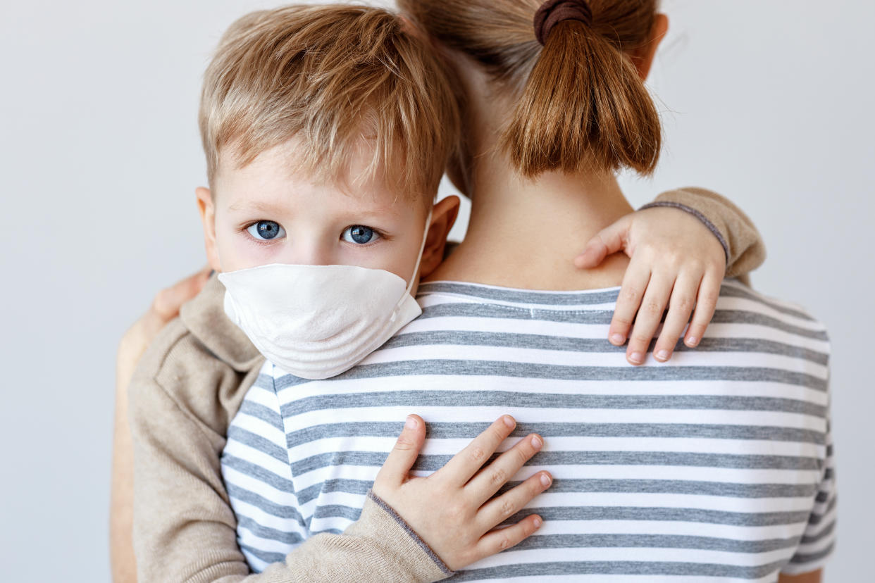 Little boy in medical mask hugging crop mother and looking at camera during coronavirus outbreak against gray background