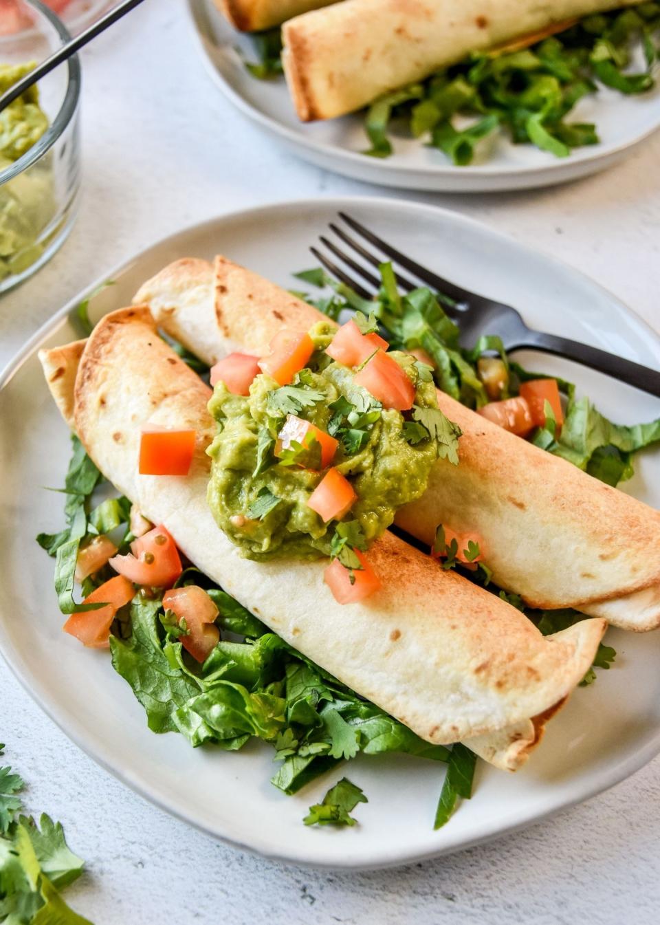 Two chicken taquitos on a plate topped with guacamole and tomatoes