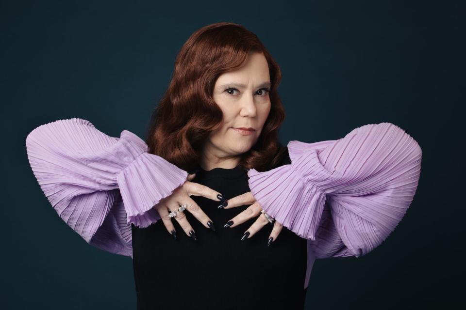 Alex Borstein in a black dress with purple sleeves, spreading her hands over her chest, elbows held high.