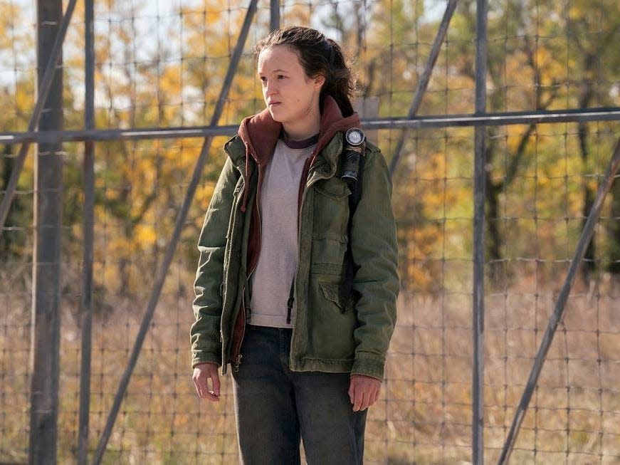 bella ramsey as ellie in the last of us, standing in front of a chain link, heavy fence in episode three