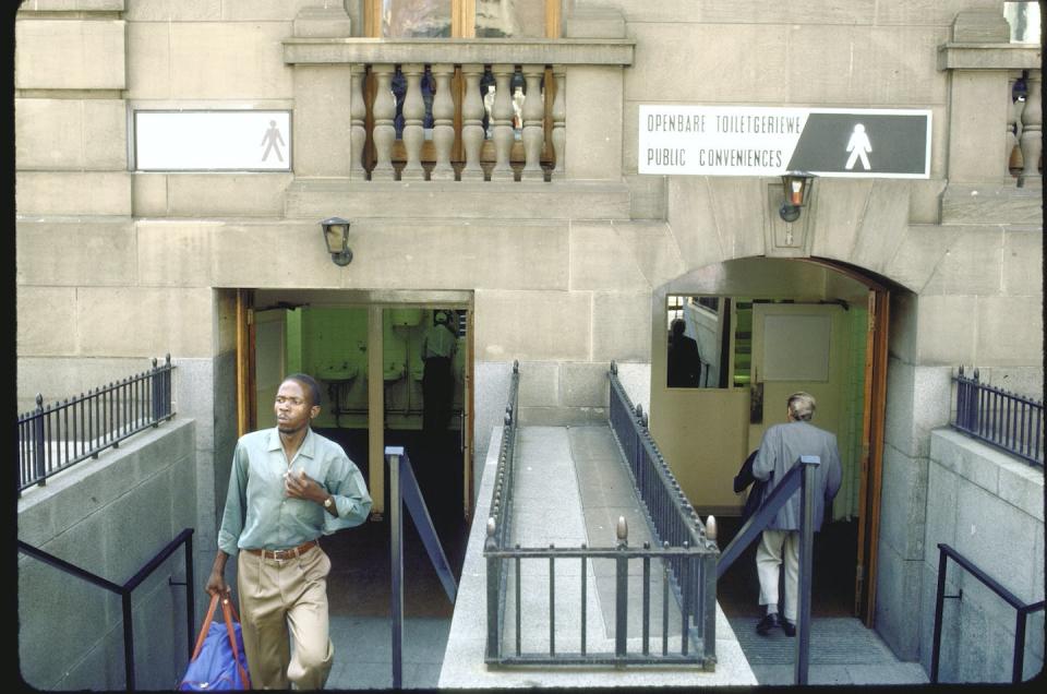 A Black man walks away from a limestone building, while a white man is seen entering on the other side. There are two signs above the entryways, one that shows a black man and the other shows a white man.