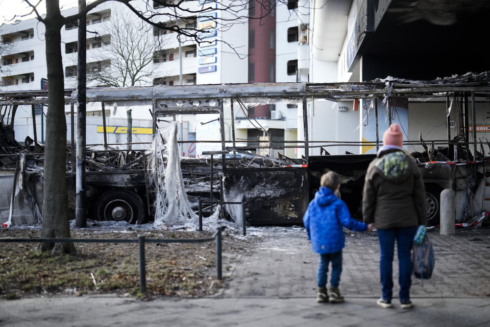 A woman with a child look to a burned-out bus standing beneath a residential building in the district Neukoelln in Berlin, Germany, Tuesday, Jan. 3, 2023. People across Germany on Saturday resumed their tradition of setting off large numbers of fireworks in public places to see in the new year. The bus was set on fire during the New Year's celebrations. (AP Photo/Markus Schreiber)