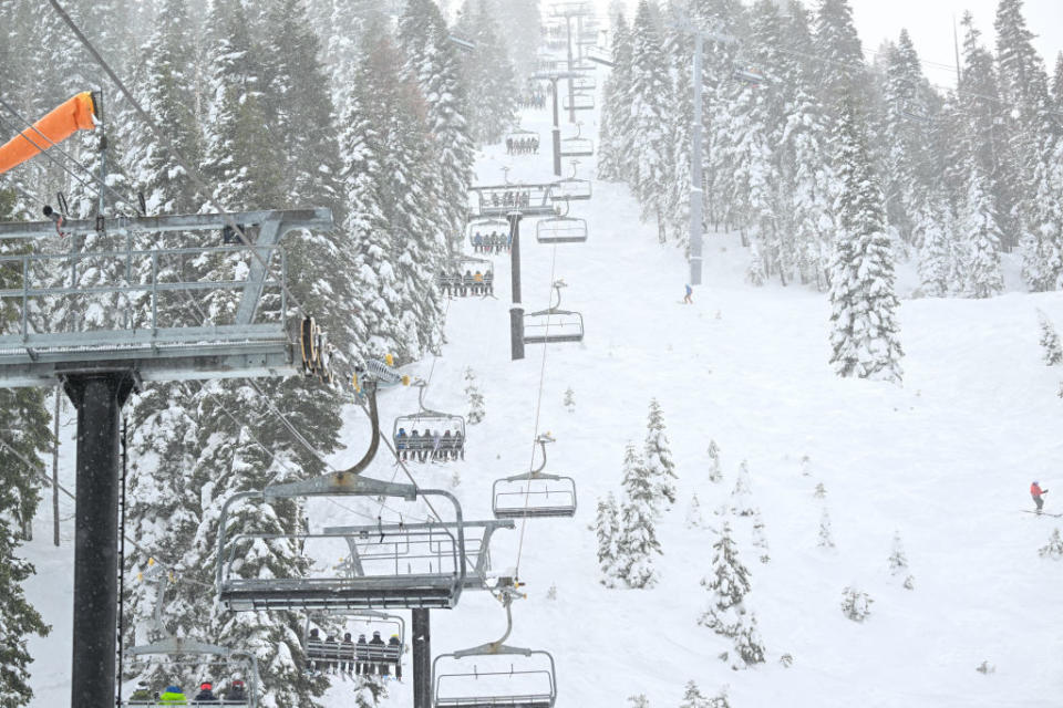 Ski lift with multiple chairs ascending, surrounded by snowy trees, skiers below
