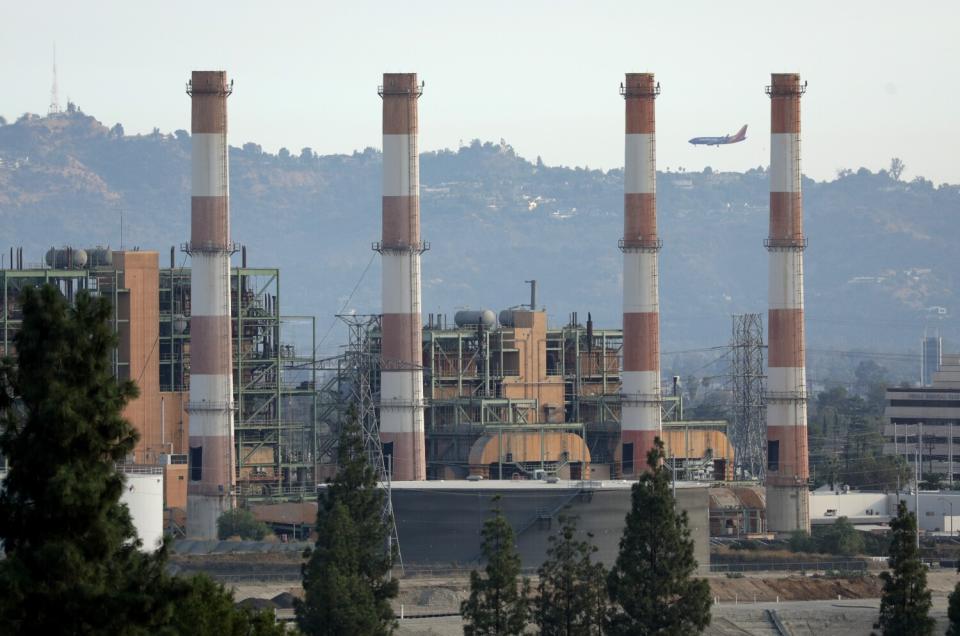 An airliner passes behind red and white striped smokestacks at a power plant