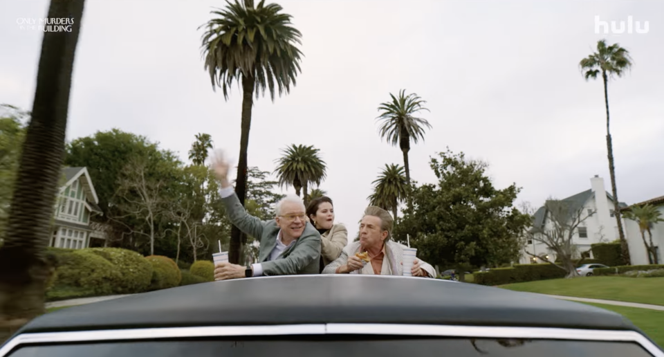 Steve Martin, Selena Gomez, and Martin Short ride in a convertible, waving and holding drinks, with a scenic suburban backdrop featuring palm trees