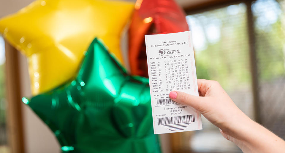 An Oz Lotto ticket is held in front of a bunch of colourful balloons.