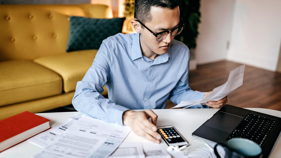 Concentrated man sitting in the living room looking at paper documents and using laptop.