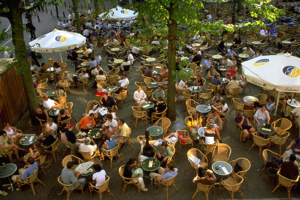 A bustling outdoor cafe with numerous people seated at tables under umbrellas, engaged in conversations and enjoying their drinks