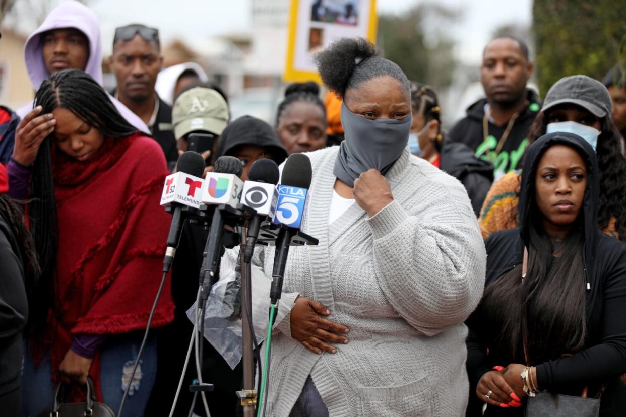 A woman speaks into a row of news microphones in front of a crowd of people