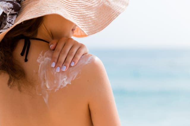 Young girl in straw hat is applying sunscreen on her back to protect her skin