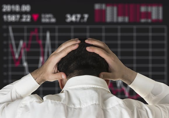 A man in a dress shirt holds his head in his hands as he stares at a declining share price chart.
