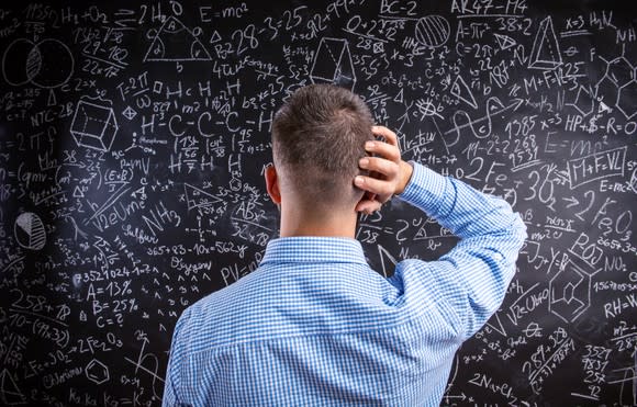 A man looking at a chalkboard full of math formulas. He has one hand placed at the side of his head.