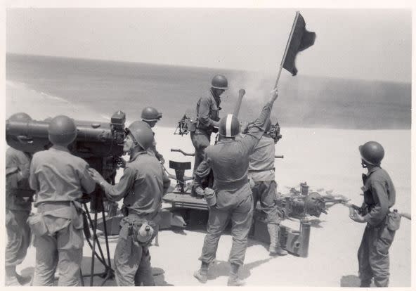 A black and white image shows people in old military uniforms holding up a flag and firing a weapon out over the ocean.