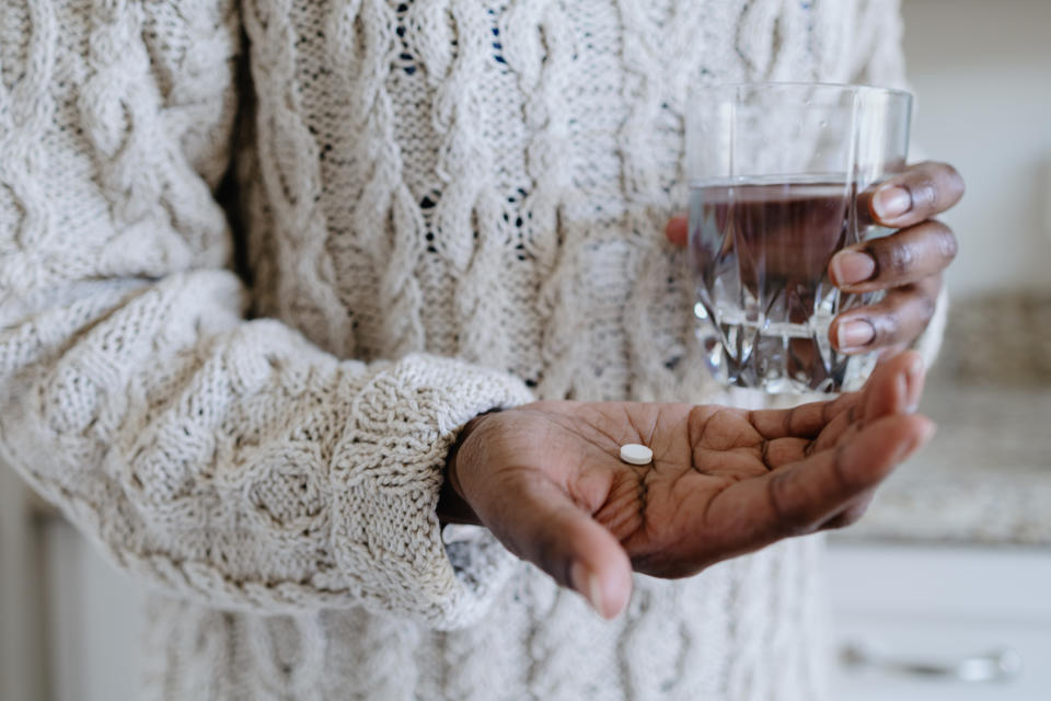 A person's hand holding a pill and a glass of water, wearing a thick sweater