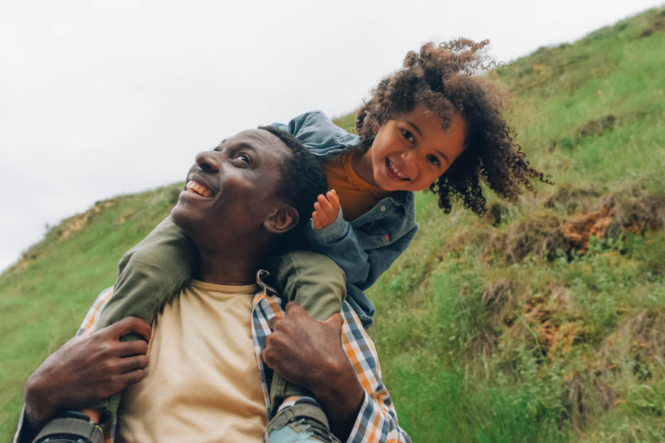 A father carries his smiling child on his shoulders outdoors on a grassy hillside. Both are happy and enjoying the moment