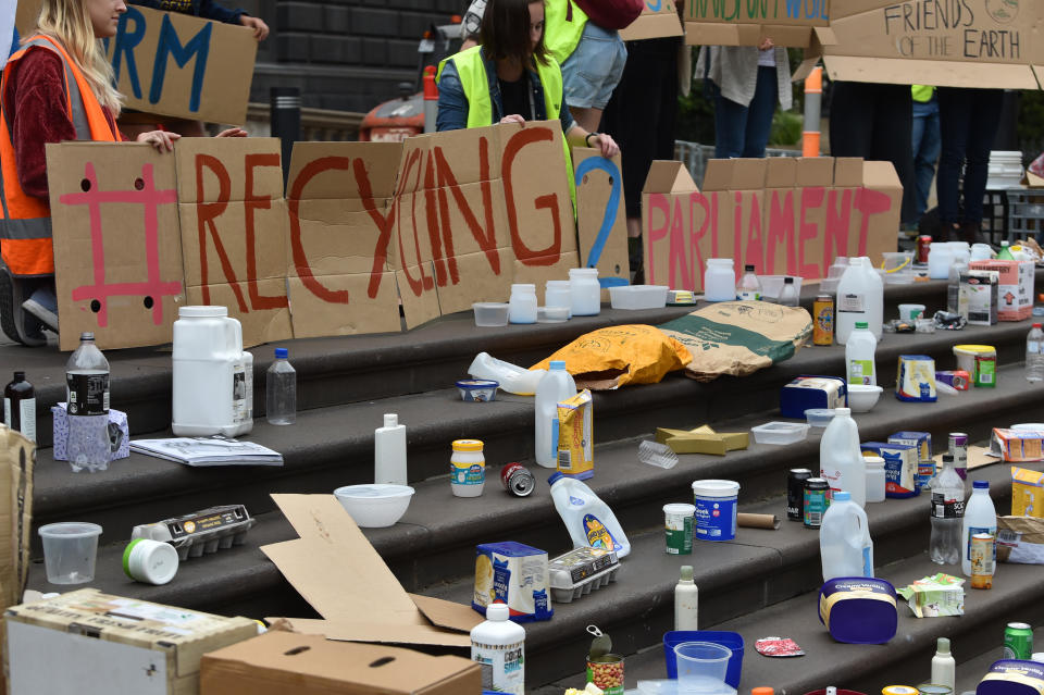 Protesters hold signs in front of plastic bottles and household recycling waste. 