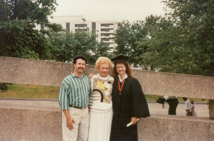 A man, an elderly woman, and a young woman in a graduation gown stand together outside, smiling for a photo