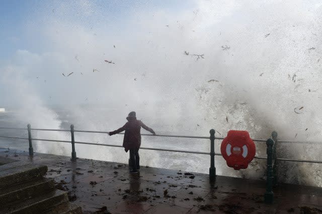 A woman walks past waves crashing on the sea wall at Penzanze, Cornwall
