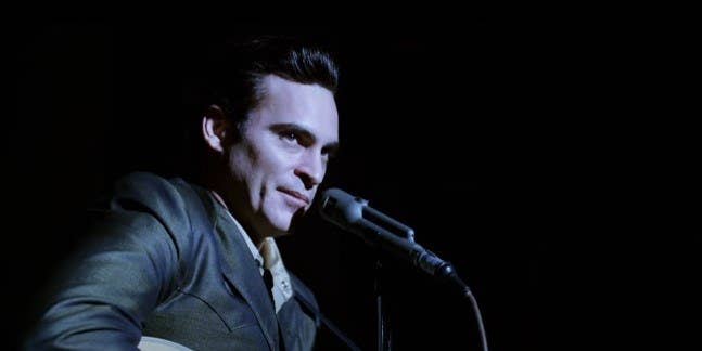 Joaquin Phoenix as Johnny Cash, wearing all black on a black background with a bright light illuminating his face as he sits in front of a microphone with a guitar