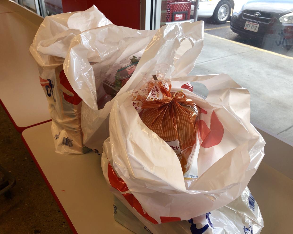 Three plastic bags of groceries on ledge in front of window at Save A Lot, window showing area for shopping carts and parking lot with cars parked