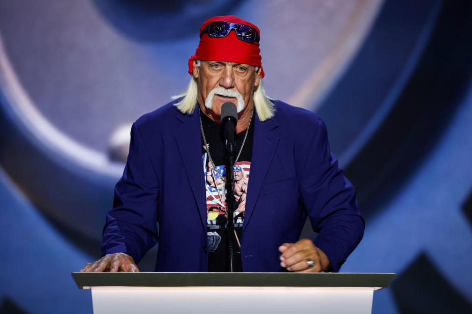 Hulk Hogan, der ein rotes Bandana und einen blauen Blazer über einem T-Shirt trägt, spricht auf einem Podium