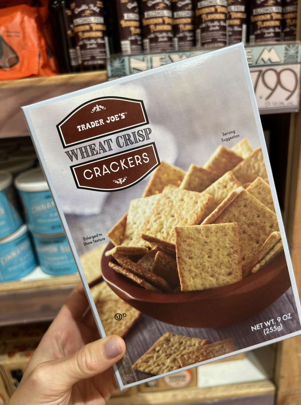 A hand holding a box of Trader Joe's Wheat Crisp Crackers in a grocery store aisle, with various other products on the shelves in the background