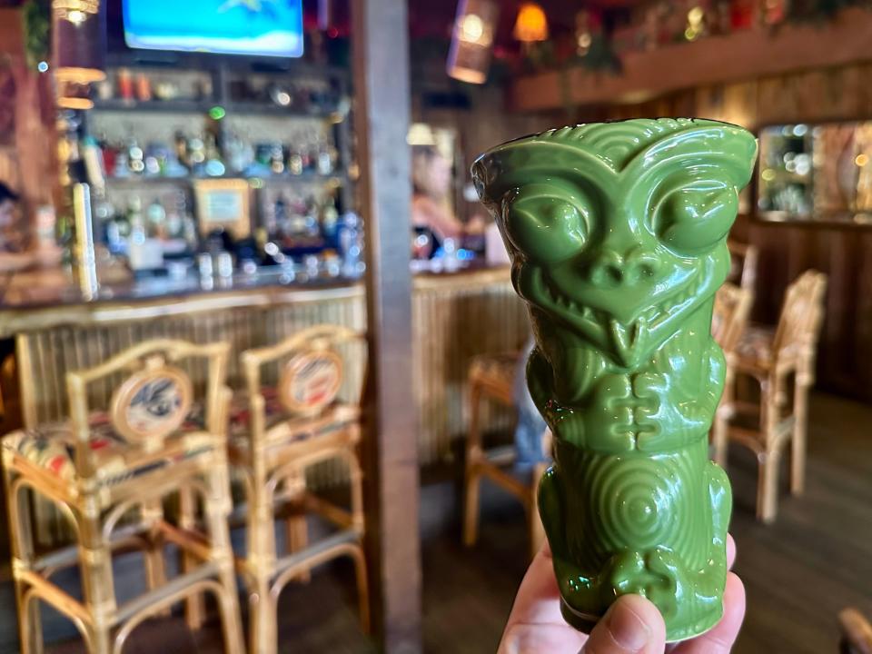 A hand holds a tiki cup and several chairs line around a bar area in the background