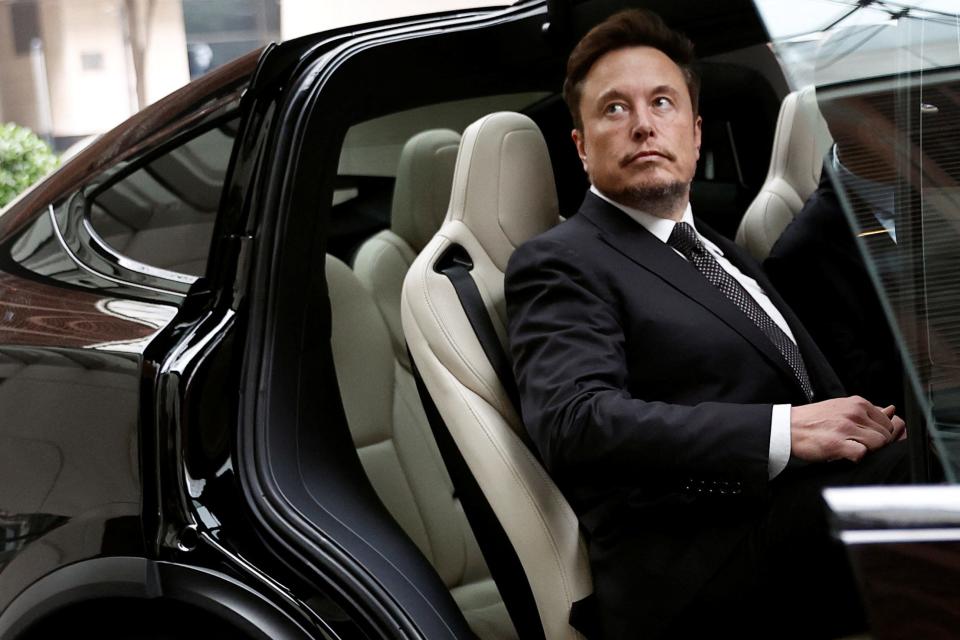 Elon Musk sitting in the a passenger seat of a black Tesla with a white interior while wearing a black suit and tie.